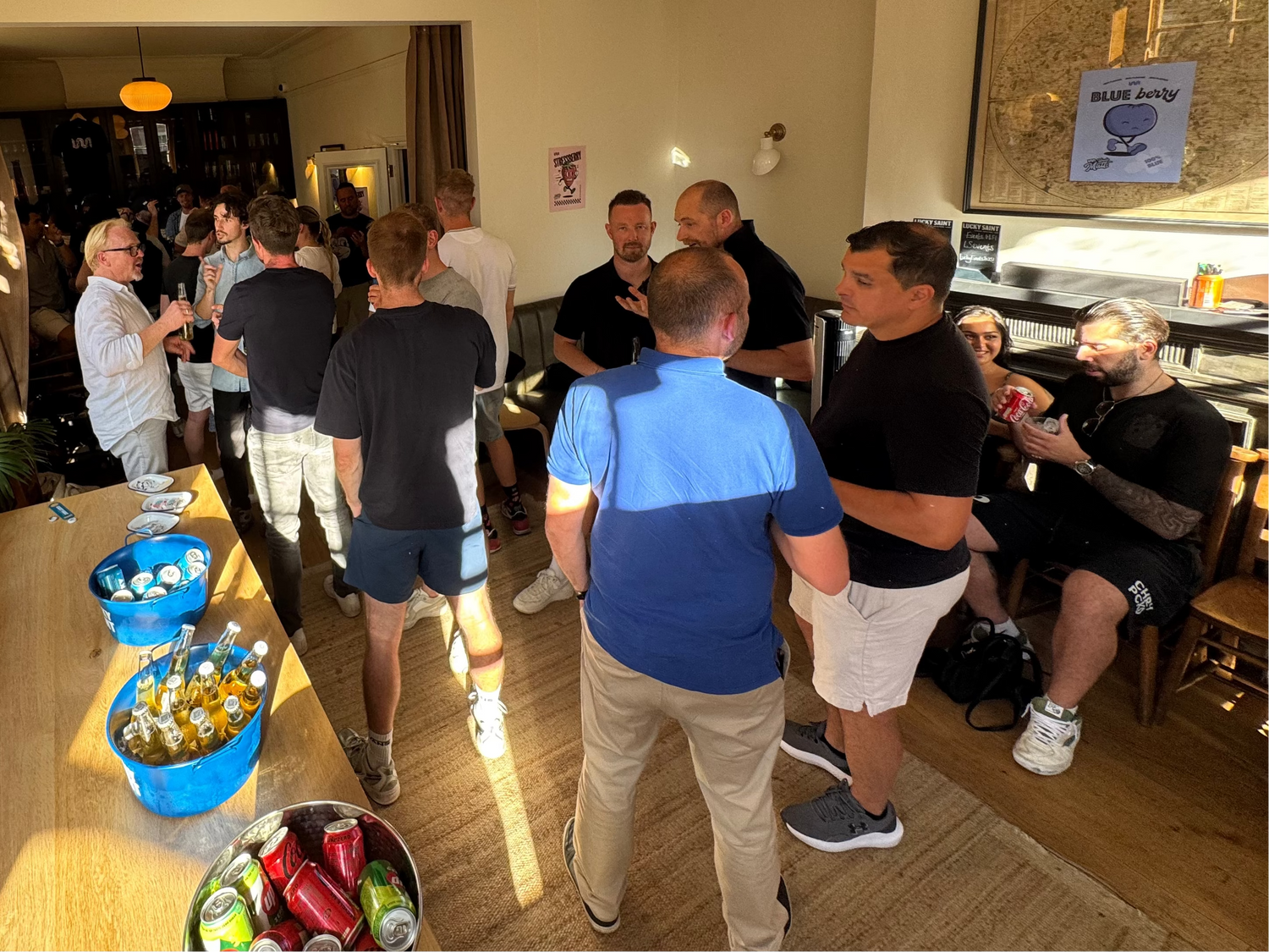 A group of men gathered in a bright, sunlit room with wooden floors and windows, standing around tables filled with snacks and hats. Several hats with red, blue, and gray branding are lined up on a table in the foreground.